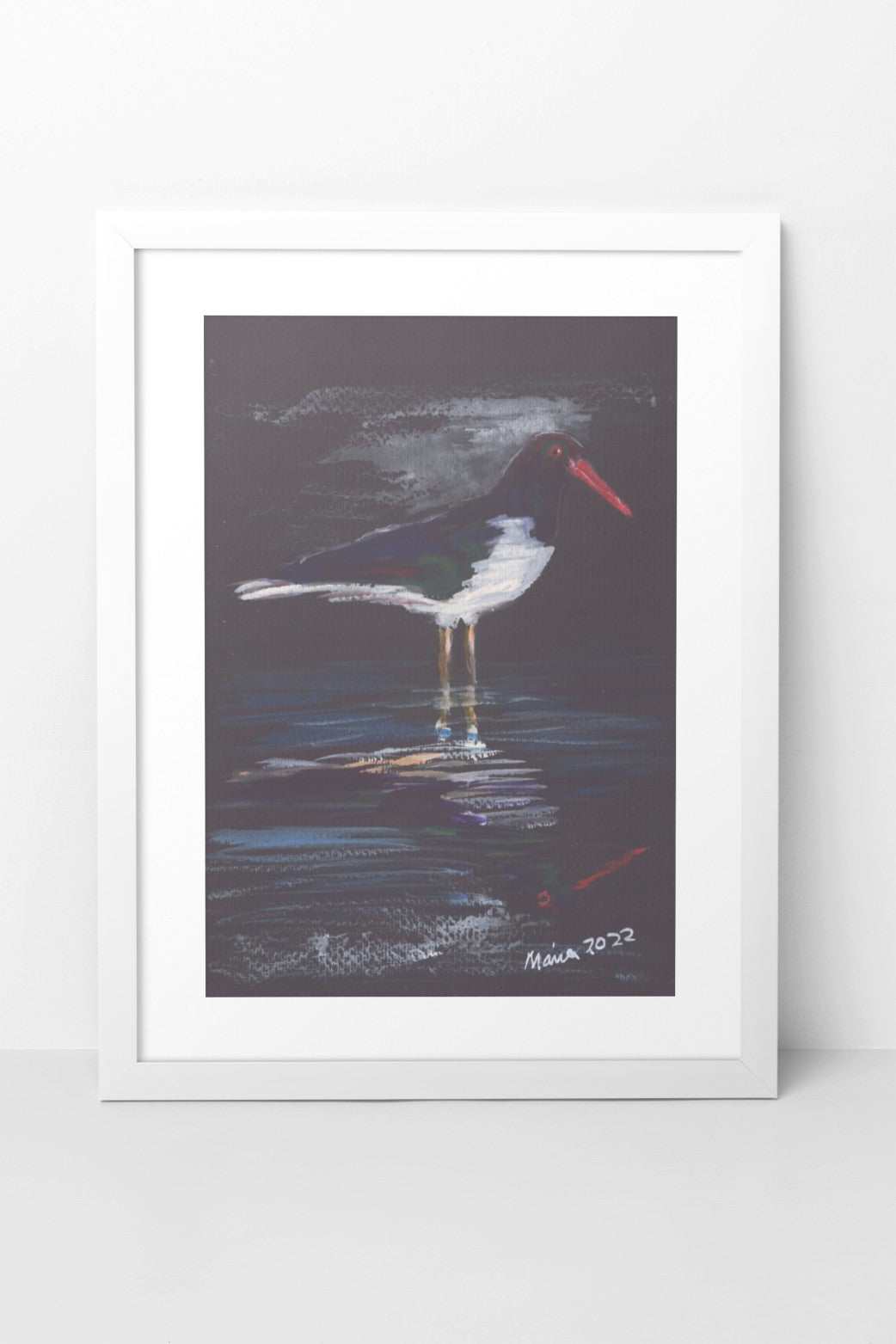 Gouache painting on black watercolor paper depicting an oystercatcher, a black-and-white bird with a bright red beak, standing in shallow water with its reflection visible in the still water. The bird's mild colors contrast with the vibrant red beak and the black paper background. The A4-sized artwork is displayed in a white mock-up frame against a neutral background, emphasizing the striking contrast and serene reflection.