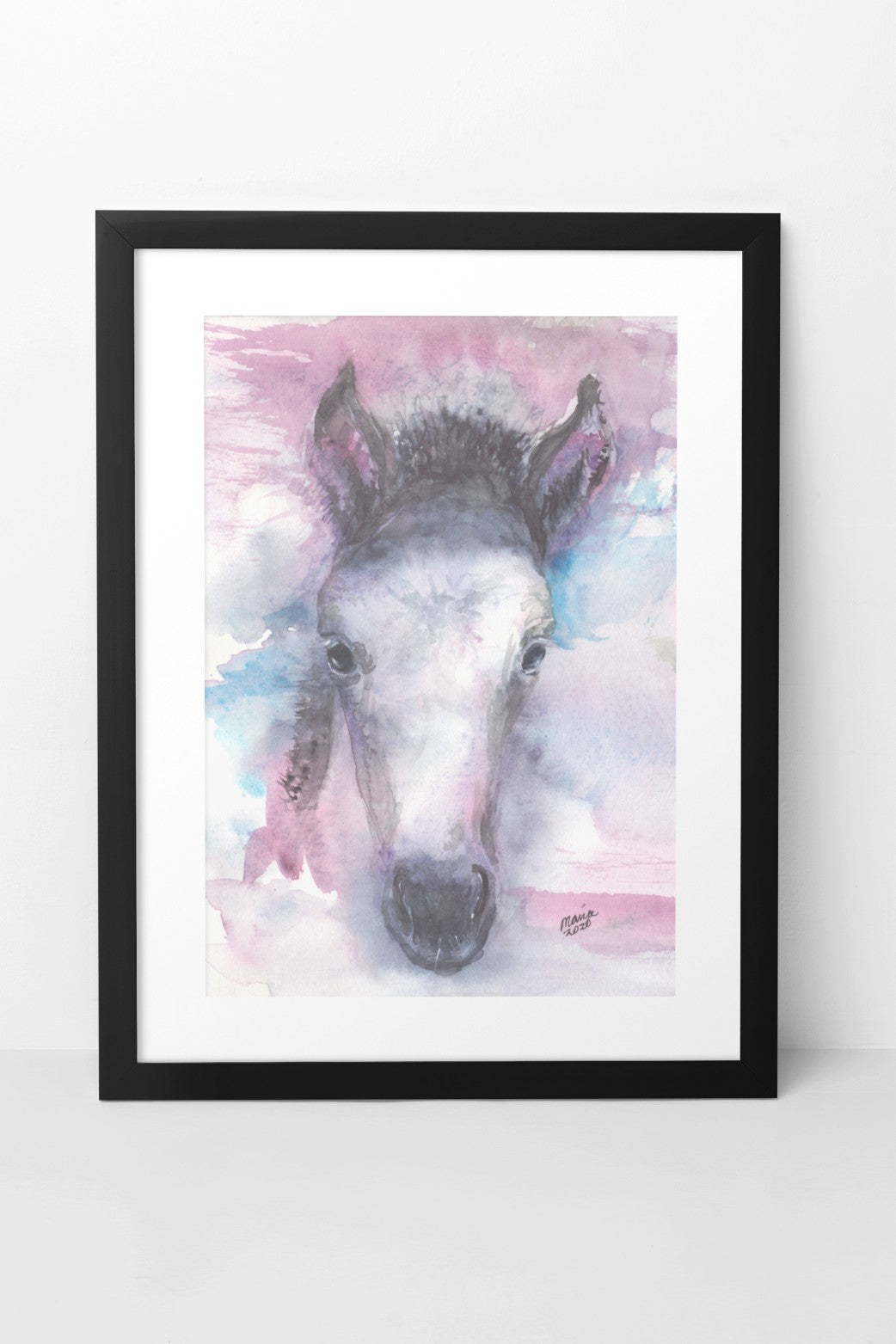 Watercolor painting of a pretty foal, depicted in soft colors with a close-up of its face. The foal has a dark mane, perky ears, and is primarily white. The colors used are a mix of pink, blue, black, and white, creating a delicate and gentle image. The A4-sized artwork is shown in a black mock-up frame against a neutral background, highlighting its soft and serene qualities.
