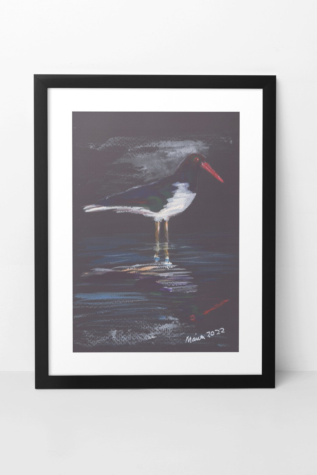 Gouache painting on black watercolor paper depicting an oystercatcher, a black-and-white bird with a bright red beak, standing in shallow water with its reflection visible in the still water. The bird's mild colors contrast with the vibrant red beak and the black paper background. The A4-sized artwork is displayed in a black mock-up frame against a neutral background, emphasizing the striking contrast and serene reflection.